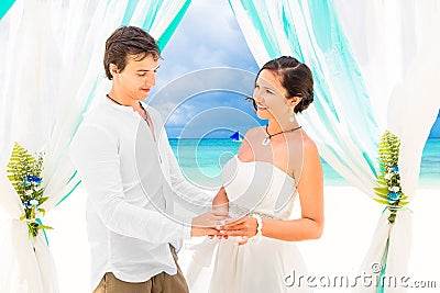 Bride giving an engagement ring to her groom under the arch decorated with flowers on the sandy beach. Wedding ceremony on Stock Photo