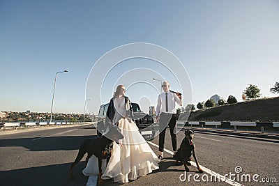 Bride and groom, in black glasses and leather jackets with Dobermans near the car on the road Stock Photo