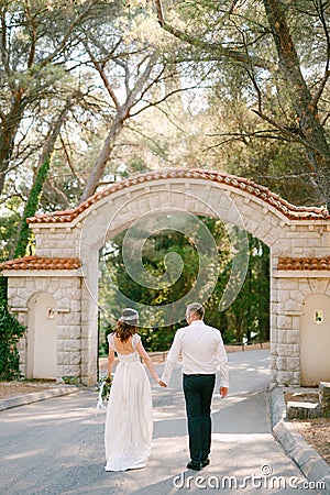 The bride and groom go to the beautiful arch at the entrance to the park holding hands, back view Stock Photo