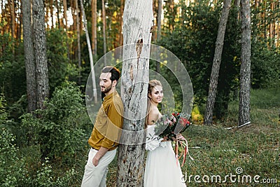 Bride and groom lean on the tree from different sides. Newlyweds are walking in the forest. Artwork Stock Photo