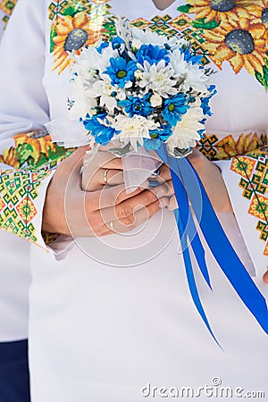 Bride and groom in shirts embroidered with sunflowers hold a bridal bouquet of blue and white chrysanthemums with blue ribbon. Stock Photo