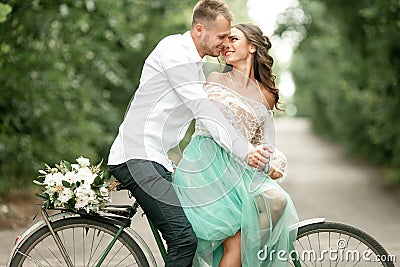 Bride and groom sit on bicycle on forest road, embrace and smile Stock Photo