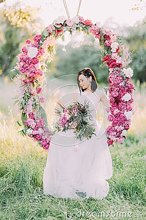 The bride with the hair accessories holding the bouquet of peonies, looking at the ground and sitting in the wedding Stock Photo