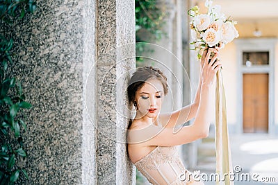 Bride lowered her eyes, leaning against a pillar with raised hands with a bouquet of flowers. Lake Como Stock Photo