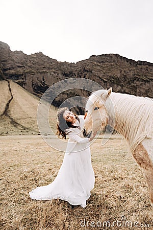 The bride strokes the face of a cream-colored horse with a luxurious mane, in a field of yellow grass, against a rocky Stock Photo