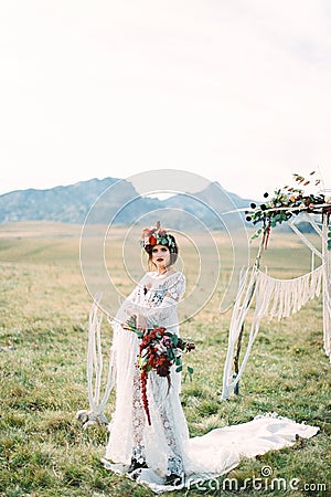 Bride in a wreath with a bouquet stands on a white carpet in front of a wedding arch in a meadow Stock Photo