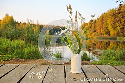 Bright autumn field bouquet of flowers in the hand-made ceramic vase at a bridge on the pond/lake. Autumn wood on the background Stock Photo