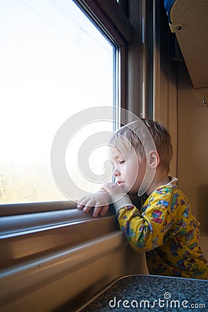 A bright, cute three-year-old boy riding a train looks out the window, behind which a dull landscape sweeps through Stock Photo