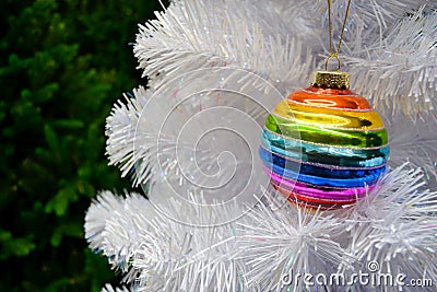 Bright glass rainbow colored Christmas ball, bauble hanging on a white artificial christmas tree. On background of green christmas Stock Photo