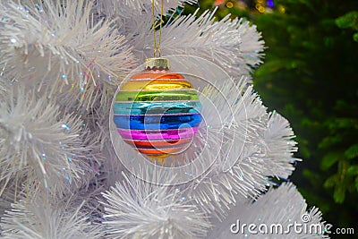 Bright glass rainbow colored Christmas ball, bauble hanging on a white artificial christmas tree. On background of green christmas Stock Photo