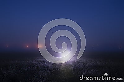 A bright mysterious light on a path on a misty night, with glowing lights on the horizon Stock Photo