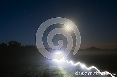 A bright mysterious light trail on a country path on a misty night, with glowing moon in the night sky Stock Photo