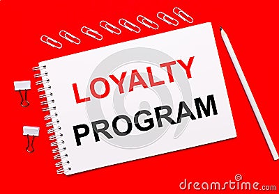 On a bright red background, a white pencil, white paper clips, and a white notebook with the text LOYALTY PROGRAM Stock Photo