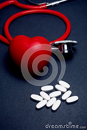 Bright red stethoscope and heart and pile of white pills on a dark blue surface, Health and treatment of heart related disease Stock Photo