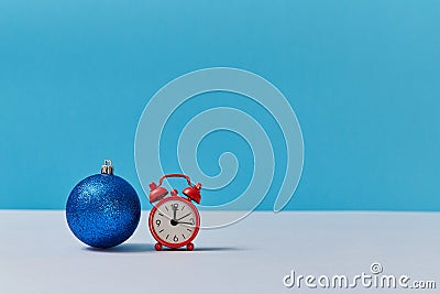 Bright shiny ball for decor. Red clock showing twelve oclock. Happy new year. Xmas concept. Copy space. Blue backdrop Stock Photo