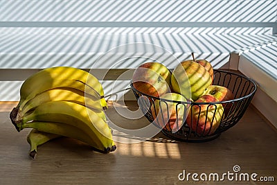 Bright sun shining through window blinds and casting stripes on bananas and aplles in the basket Stock Photo