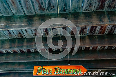 Brightly colored Male Bathroom arrow sign in all caps/capital letters hanging on chain suspended from ceiling. Stock Photo