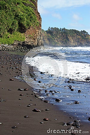 Brightly colored rocks strewn along a black sand beach ending in a cliffside and meeting the Pacific Ocean in Hawaii Stock Photo