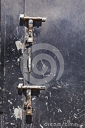 Old scratched black steel door with two large rusty latches and padlocks hanging from them. Stock Photo