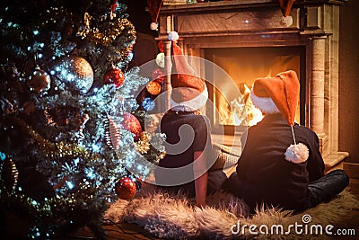 Back view, brother and sister wearing Santa`s hats warming next to a fireplace in a living room decorated for Christmas. Stock Photo