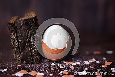 Brown boiled half-peeled egg and pieces of black bread on a dark wooden background with scattered salt and shell Stock Photo