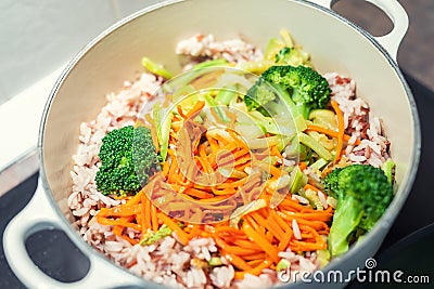 Brown rice with organic unmixed peeled sliced vegetables cooking in cast iron saucepan. Bright broccoli, carrot and Stock Photo