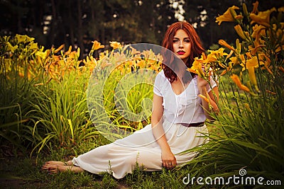 Brunette caucasian woman in white dress at the park in red and yellow flowers on a summer sunset holding flowers sitting on the gr Stock Photo