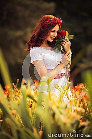 Brunette caucasian woman in white dress at the park in red and yellow flowers on a summer sunset holding roses Stock Photo