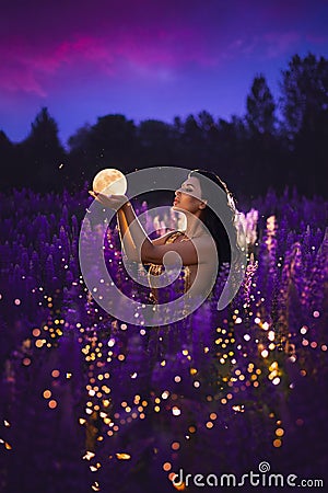 A brunette girl holding a moon in her hands and standing among a blooming purple lupine field Stock Photo