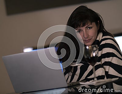 Brunette girl is sitting in the at home behind a laptop, smiling and looking at the camera with a glass of wine Stock Photo