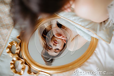 Brunette lying on bed and looking at gold-rimmed mirror. Bride holding pillow under hands and admiring reflection. Stock Photo