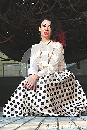 brunette with red hair in a beige blouse and skirt with black polka dots is leaning against a leafless tree Stock Photo