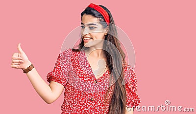 Brunette teenager girl wearing summer dress looking proud, smiling doing thumbs up gesture to the side Stock Photo