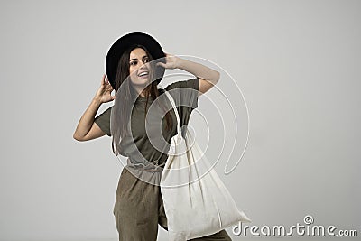 Brunette woman in green t-shirt and black hat holding cotton shopper bag with vegetables, products in white room. Eco Stock Photo