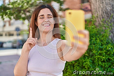 Brunette woman taking a selfie photo with smartphone smiling with an idea or question pointing finger with happy face, number one Stock Photo