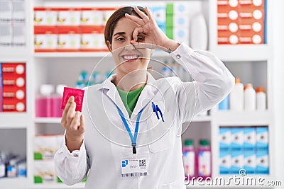 Brunette woman working at pharmacy drugstore holding condom smiling happy doing ok sign with hand on eye looking through fingers Stock Photo