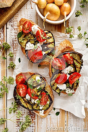 Bruschetta with grilled eggplant, cherry tomatoes, feta cheese, capers and fresh aromatic herbs on a wooden table. Delicious Medit Stock Photo