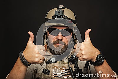 Brutal man in military desert uniform and body armor shows two fingers up on black background in Studio. The bearded player in the Stock Photo