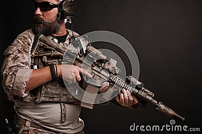 Brutal man in the military desert uniform and body armor stands in a fighting rack and holds his rifle on a black background. The Stock Photo