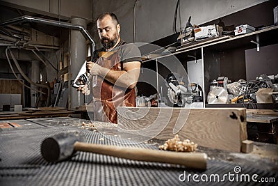 Brutal master carpenter in an apron adjusts plane in workshop Stock Photo