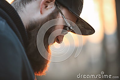 A brutal young man with a huge beard in sunglasses and a cap in the woods at sunset. Stock Photo