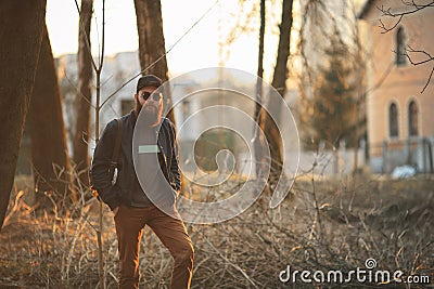 A brutal young man with a huge beard in sunglasses and a cap in the woods at sunset. Stock Photo