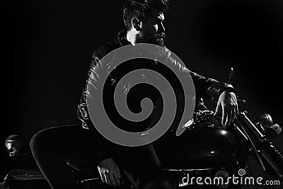 Brutality and masculine concept. Man with beard, biker in leather jacket lean on motor bike in darkness, black Stock Photo