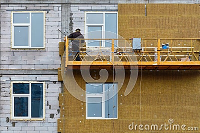 A builder standing on a suspended platform insulates the facade of a high-rise building under construction with mineral Stock Photo