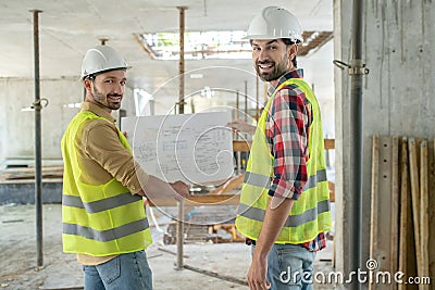 Building workers in yellow vests turning thier heads to camera, holding blueprint Stock Photo