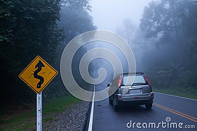 A bullet riddled curvy road sign and silver SUV car on the asphalt road through a misty mysterious tropical forest. Somewhere in Stock Photo