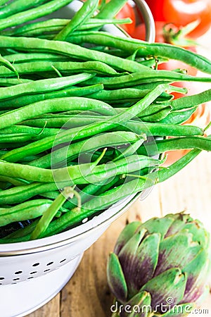 Bunch of fresh raw green beans in white metal colander on plank wood kitchen table, tomatoes, artichokes Stock Photo