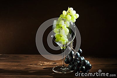 A bunch of green grapes and a wine glass on a dark background in the conditions of artificial hard lighting close up. In a glass o Stock Photo