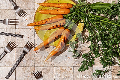 Bunch of grilled carrot roots with green leaves and salt flakes on a green craft plate on stone tales of earth tones background wi Stock Photo