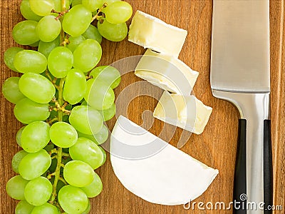 Bunch of sweet green grapes, pieces of tasty cheese camembert and chef knife on a brown wooden cutting board. Soft cheese covered Stock Photo